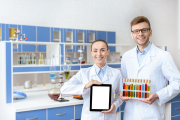 Fototapeta na wymiar Young smiling chemists in lab coats holding digital tablet and test tubes