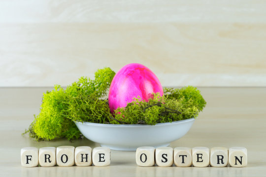 Ein kleines Osternest mit Moos und einem pink farbenen Osterei, davor die Worte Frohe Ostern aus Buchstabenw&uuml;rfeln gelegt, Holzhintergrund und Textfreiraum