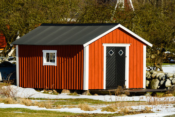 Red, white and black wooden shed in early spring with some snow still visible on the ground.