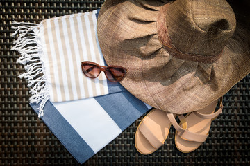 The concept of flatlay summer accessories of white, blue and beige Turkish towel, sunglasses, beige leather ladies sandals and straw hat on a rattan lounger.