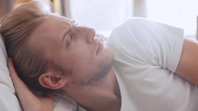 Upset young handsome Caucasian male wearing white shirt lying in upset in bed in the morning with girfriend next to him in slowmotion