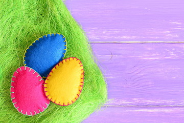Pink, yellow and blue felt Easter eggs in a grass of green sisal fibre and on purple wooden background with copy space for text. Vivid Easter background. Closeup