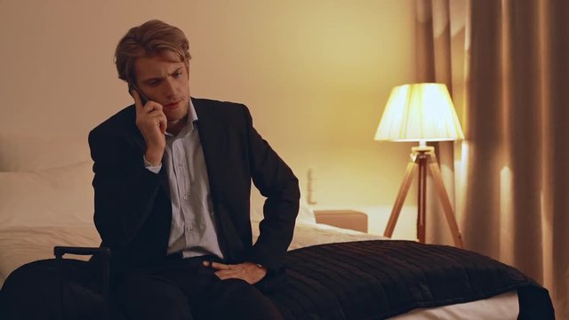 Concentrated younf Caucasian busniess man in suit sitting at the edge of bed and talking on mobile phone in slowmotion