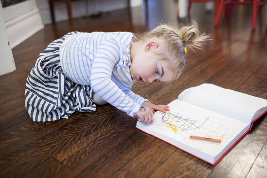 Young girl doodling in her sketchbook with crayons on the dining room floor; Toronto, Ontario, Canada
