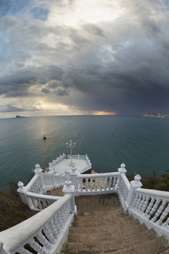 Steps leading down to the turquoise water with the sun glowing on the horizon under storm clouds; Benidorm, Spain