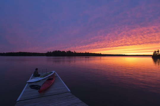 A young woman sits at the end of the dock with kayaks watching the sun set over a lake; Ontario, Canada