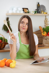 Young  brunette woman is looking a new recipe for cooking at the kitchen. Housewife holding wooden spoon and tablet computer while searching internet. Food and health concept.