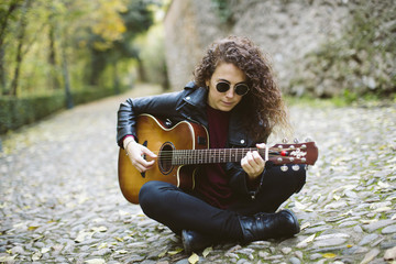 Beautiful young woman playing guitar on forest. Girl wearing black jacket and sunglasses, fashion...