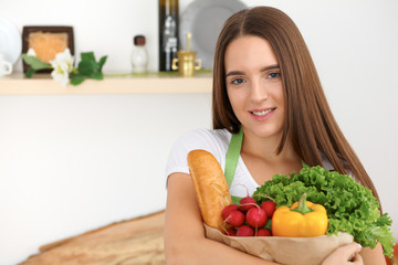 Young caucasian woman in a green apron is holding paper bag full of vegetables and fruits while smiling in kitchen