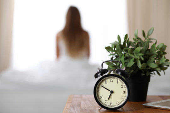 Alarm clock standing on bedside table has already rung early morning to wake up woman in bed sitting in background. Early awakening, not getting enough sleep, oversleep, time line concept