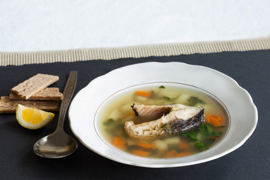 Fish soup on black stone background. Vintage white plate with sliced fish, potato, carrot and herbs. slice lemon, aged spoon, bread. Up view.