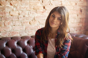 Happy smiling latin girl sit on the red leather sofa and look to the camera. Brunette attractive woman in plaid shirt with red brick wall background under sun from window. Brightening effect.