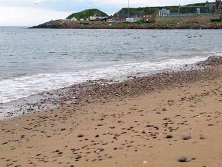 Sandstrand von Stonehaven in Schottland mit bunten Steinen. Im Hintergrund ist ein Teil des alten Fischerdorfes zu sehen.