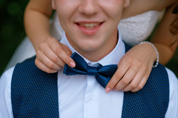 Bride corrects a blue bow tie groom, who smiles in a white shirt and waistcoat snem summer day in the park