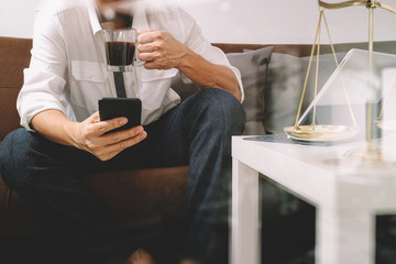 Justice and Law context.Male lawyer hand sitting on sofa and working with smart phone,digital tablet computer docking keyboard with gavel and document on living table at home,filter effect