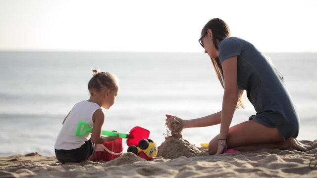 Young mother with little doughter playing with sand on the beach