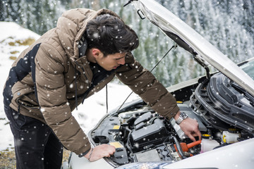 Young man near car with open hood inspecting engine in winter. Snowy forest on background