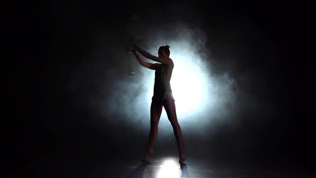 Girl gymnast with mace in hand revolve around him. Black background. Light rear. Silhouette. Slow motion