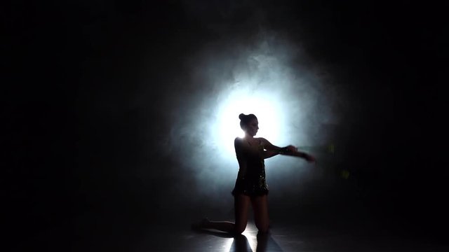 Rhythmic gymnast throws mace up and catches her. Black background. Light rear. Silhouette. Slow motion