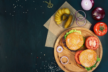 Craft beef burgers on round wooden cutting board with vegetables. Flat lay on black textured background.