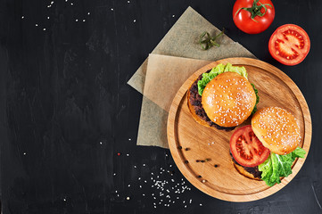 Craft beef burgers on round wooden cutting board with vegetables. Flat lay on black textured background.