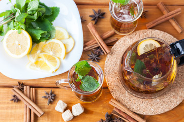 Two clear glasses and teapot of hot black herbal tea with lemon, mint and Melissa leaves on light rustic wooden table. Summer, Autumn, winter drink. Cinnamon and cane sugar for decoration. Top view.