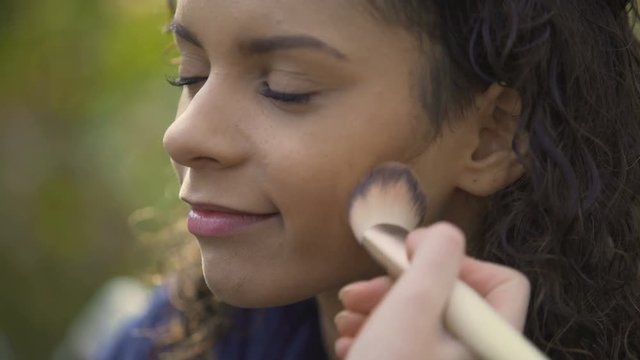 Professional makeup artist evens up model skin tone with brush, movie shooting