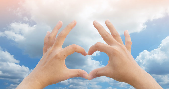 hands doing heart symbol have blue sky with white cloud with orange light is background