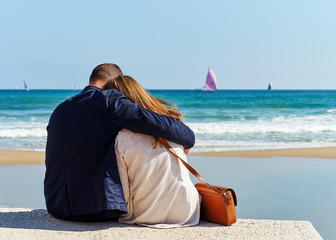 Love story on the beach. Woman and man sitting embracing and looking and admiring the sea. Running athlete on the sea shore