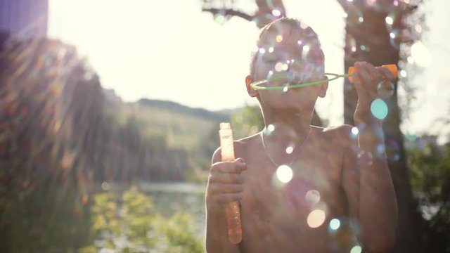 Young handsome happy boy blowing bubbles outdoors on sunny day in summer on sunshine background with blurred magic bokeh. 1920x1080