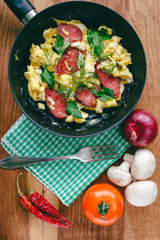 Fresh cooked scrambled eggs in pan with sausage and herbs. fork, vegetables, napkin on wooden board, top view.