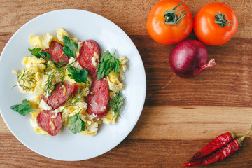 Fresh cooked scrambled eggs with sausage and herbs in white plate. Tomato, chilli pepper, onion on wooden board top view