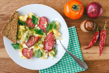Fresh cooked scrambled eggs with sausage and herbs in white plate. Tomato, chilli pepper, onion on wooden board top view