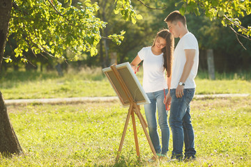 Young couple drawing at easel by colorful variegated paints. Pretty young woman and handsome guy having fun with paints outdoors.