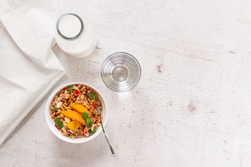 Healthy breakfast - plate of granola with orange slices, mint and seeds, bottle of milk, water, copy space on white wooden table
