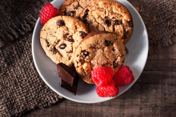 Chocolate chip cookies on white plate dark old wooden table with red raspberry and  place for text.,  freshly baked. Selective Focus with Copy space..