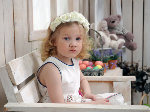 Beautiful little girl in white dress and wreath.The wooden walls of the room, the window. Basket with colored Easter eggs