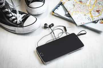 Essential travel accessories: hipster sneakers, maps, telephone with text and headphones on wooden background. Selective focus. Traveling concept