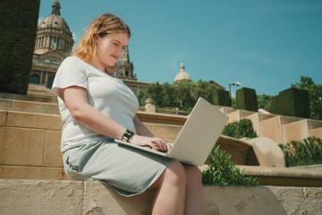 Plus size beautiful model girl working on her laptop while sitting on stairs in sunny day, young woman in white t-shirt and grey skirt is having online chat with her business colleagues using net-book