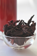 Hibiscus petals in a glass bowl and hibiscus infusion tea in the background. Deep red herbal hot beverage. Isolated on white


