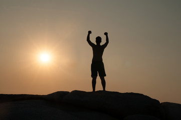 Positive male holding hand up and expressing gladness while standing on stone in Sunset.