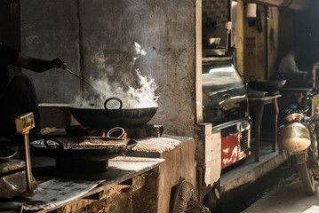 Unrecognizable man cooking in fatiscent big pan or wok in a small street food stall. White smoke coming out from the pan, hand and arm only visible. Street food in India.