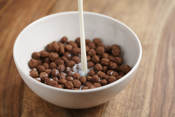 pouring milk into chocolate cereal balls in white bowl for breakfast on wooden table