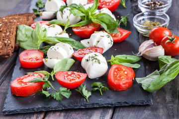Cherry tomatoes, mozzarella cheese, basil, pesto sauce, dark rye grain bread and spices on black slate stone chalkboard. Italian traditional caprese salad ingredients. Mediterranean food. 
