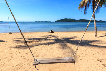 A wooden swing seat hanging on coconut tree by the sea with nice clean beach and deep blue sky.