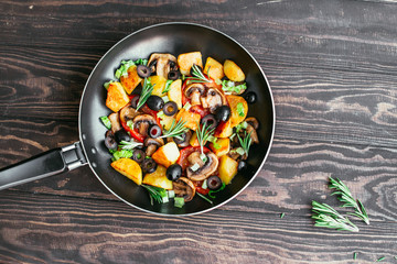 fried potatoes in a pan with tomatoes, mushrooms, olives, greens decorated with sprigs rosemary on a wooden table vegetarian food Top view