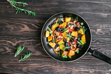fried potatoes in a pan with tomatoes, mushrooms, olives, greens decorated with sprigs rosemary on a wooden table vegetarian food Top view