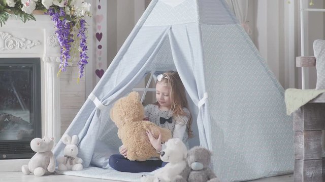 Girl throws up a teddy bear. Little cute girl with blond hair sitting in a toy house surrounded by soft toys. Playroom for kids with Teepee or DIY tent. Wigwam for children in a room.