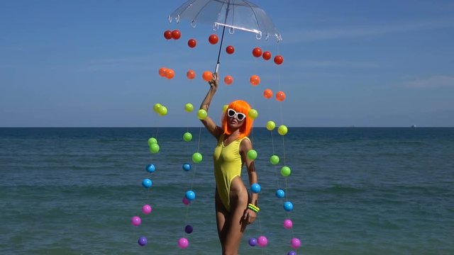 Sexy woman posing with coloured balls attached to umbrella during summer day over sea and sky background. Creative look of woman in yellow swimsuit, orange wig and white sunglasses