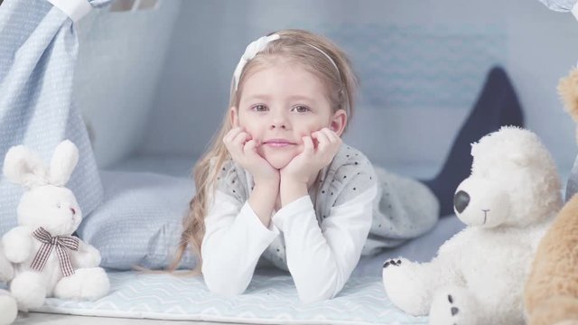 Little cute girl lying on the floor in the nursery in tepee tent and put her hands on her cheeks. Teddy bear soft about girls. Little girl sword and dreams, lying in tepee tent.